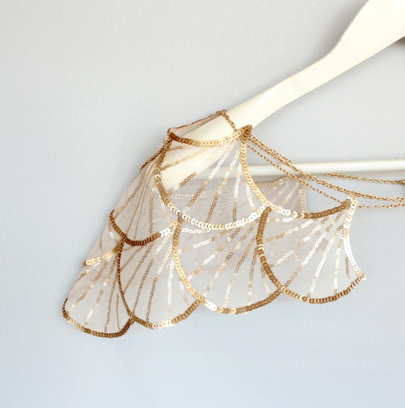 Gold sequin embroidered wing-shaped bridal shoulder capelet displayed on a white hanger, showing the decorative chain drapes and scalloped tulle edges.