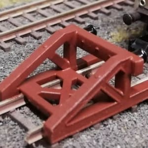 Può includere: Un respingente per treni modello color amaranto, progettato per fermare i treni modello, è posizionato su un binario ferroviario in miniatura. Il respingente ha un design geometrico triangolare. Il binario è posato su un letto di ghiaia grigia.