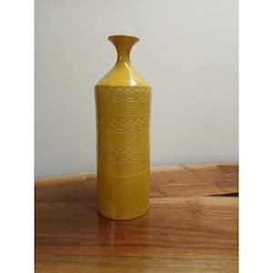 May include: A tall, mustard-yellow ceramic vase with a slender neck and a wide base. The cylindrical body features a repeating geometric pattern of rectangles and diamonds. The vase is approximately 12 inches tall.