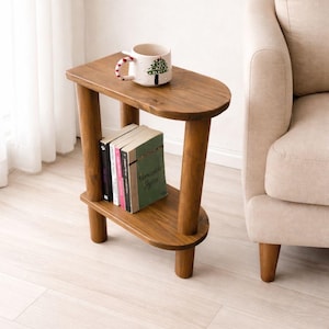 May include: A wooden side table with a curved design, featuring two shelves and four rounded legs. A white mug with a Christmas tree design sits on top, and several books are on the lower shelf. The table is approximately 61 cm tall.
