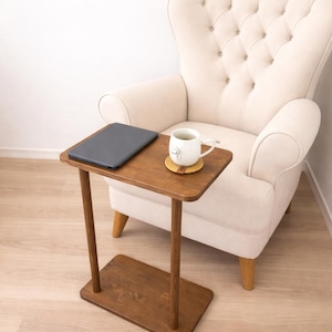 Puede incluir: Pequeña mesa auxiliar rectangular de madera oscura. Un portátil y una taza blanca con un platillo están sobre la mesa. La mesa está junto a un sillón color crema.