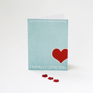 May include: A light blue card with the text "Just in case you were wondering, I totally love you" and a red heart. The card is on a white surface with red confetti.