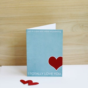 May include: A light blue card with the text "just in case you were wondering I totally love you" and a red heart. Two red paper hearts are on a white surface below the card.