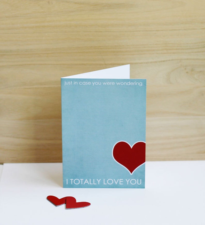 May include: A light blue card with the text "just in case you were wondering I totally love you" and a red heart. Two red paper hearts are on a white surface below the card.