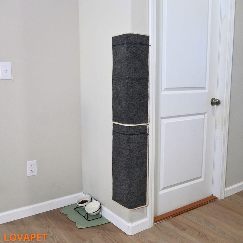May include: A dark gray cat scratcher mounted on a corner wall, with a white trim and a zipper. Below, a cat food and water bowl set on a green mat. The brand name "LOVAPET" is visible in the bottom left corner.