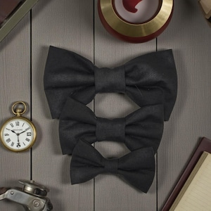 Può includere: Tre papillon neri di diverse dimensioni disposti su una superficie di legno grigio. Un orologio da tasca dorato e altri accessori vintage sono sullo sfondo. I papillon sono realizzati in tessuto strutturato.