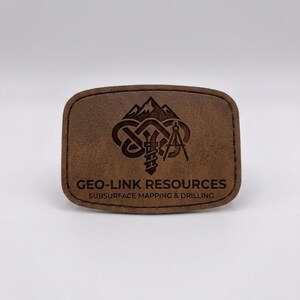 May include: A rectangular brown leather patch with rounded corners and stitched edges. The patch features a laser-engraved logo of mountains, a drill bit, and a compass, with the text GEO-LINK RESOURCES SUBSURFACE MAPPING & DRILLING below.