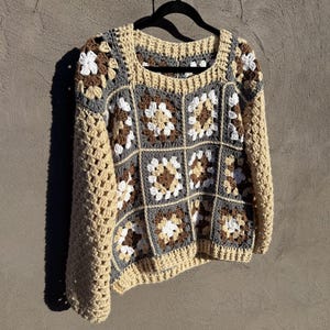 Könnte beinhalten: Ein handgefertigter Häkelpullover im Granny-Square-Design. Der Pullover hat einen quadratischen Ausschnitt und lange Ärmel. Die Farbpalette umfasst Beige, Grau, Braun und Weiß, wodurch ein Patchwork-Effekt entsteht. Der Pullover hängt an einem schwarzen Kleiderbügel.