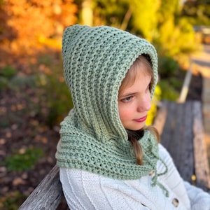 Może przedstawiać: Jasnozielony kaptur zrobiony na szydełku ze sznurkiem. Kaptur jest wykonany z teksturowanej przędzy i jest noszony na białym swetrze. Tło jest rozmyte, z jesiennymi kolorami.