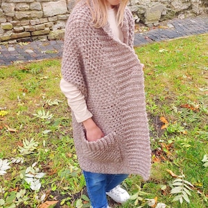 May include: A light brown, chunky knit shawl with two front pockets. The shawl is worn over a cream-colored long-sleeved shirt and blue jeans. The shawl is made of a soft, textured yarn.