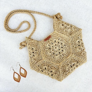 Könnte beinhalten: Handgefertigte, sechseckige, beige Häkeltasche mit einem Seilriemen und passenden Ohrringen. Die Tasche hat ein detailliertes Lochmuster und ein kleines braunes Lederetikett. Die Ohrringe sind braun mit einem dekorativen Muster.