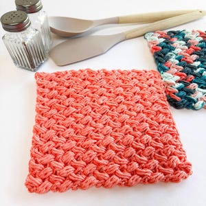 Peut inclure: Un carré de tissu éponge fait à la main, de couleur corail, avec un motif tissé texturé. Deux autres débarbouillettes en bleu sarcelle, rose et blanc sont également visibles. Des ustensiles de cuisine, dont une paire de spatules à manche en bois et des salières et poivrières, sont en arrière-plan.