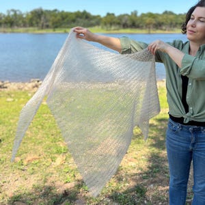 Puede incluir: Un chal de tejido abierto de color beige claro se muestra contra un fondo de un lago y árboles. El chal tiene forma triangular y una textura delicada y aireada, lo que sugiere que está hecho a mano. La tela parece suave y ligera.