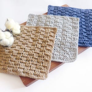 Könnte beinhalten: Drei gehäkelte Waschlappen in Beige, Grau und Blau auf einem Holzschneidebrett angeordnet. Die Waschlappen haben ein strukturiertes, gewebtes Muster. Flauschige Baumwollkugeln sind auf der linken Seite des Schneidebretts platziert.