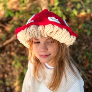May include: A handmade crochet mushroom hat. The hat has a red cap with white spots and a cream-coloured brim. The design is intended to resemble a mushroom. A whimsical accessory.