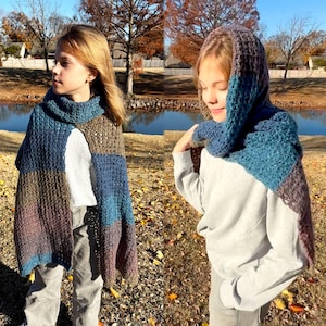 May include: Two children wearing handmade crochet capes and hoods. The capes feature a patchwork design in shades of blue, brown, and purple. The hoods are also crocheted in similar colors. The background includes a pond and trees.