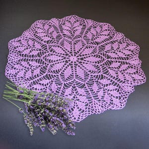 May include: A lilac-coloured, handmade crochet doily with a detailed floral pattern. The round doily features an intricate openwork design. A bundle of fresh lavender sprigs rests beside the doily, adding a natural element to the composition.