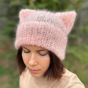 Könnte beinhalten: Ein hellrosa gestrickter Hut mit Katzenohren. Der Hut ist aus einem weichen, flauschigen Material gefertigt. Der Hut hat einen gerippten Bund. Die Katzenohren sind spitz und stehen oben heraus. Der Hut wird von einer Person getragen.