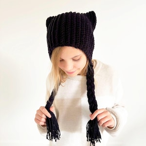 Könnte beinhalten: Eine dunkelviolette Strickmütze mit Katzenohren und langen geflochtenen Zöpfen. Die Mütze ist aus dickem, strukturiertem Garn gefertigt. Die Zöpfe hängen auf beiden Seiten des Gesichts herunter. Die Mütze wird von einer Person mit blonden Haaren getragen.