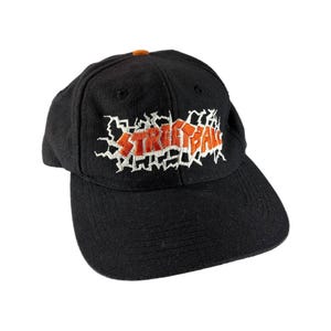 May include: Black baseball cap with the embroidered word "STREETBALL" in orange and white. The cap has a curved brim and a small orange button on top. The hat is made of a textured fabric.