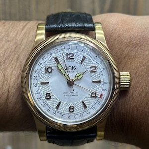 Puede incluir: Un reloj de pulsera automático Oris con caja color bronce, esfera plateada guilloché y correa de cuero negro. El reloj cuenta con una aguja indicadora de fecha, números arábigos en posiciones pares y el texto Oris, Automatic, Swiss Made.