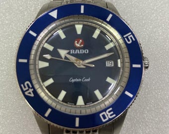 May include: A stainless steel Rado Captain Cook watch with a blue bezel and a dark blue dial. The watch features white hour markers, a date display at the 3 o'clock position, and a stainless steel bracelet. The watch has the text "RADO" and "Captain Cook" on the dial.