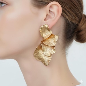 May include: Gold-coloured statement earrings with a layered, petal-like design. The earrings have a textured surface and a flowing, organic shape. The earrings are large and eye-catching, suitable for special occasions.