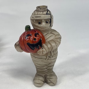 May include: A small figurine of a mummy wrapped in beige bandages, featuring dark eyes and a neutral expression. The mummy is holding a bright orange pumpkin with a carved, smiling face, featuring a green stem and black painted eyes and mouth.