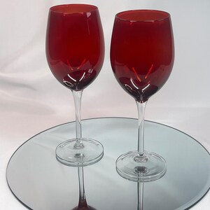 May include: Two wine glasses featuring deep red glass bowls and clear glass stems and bases. The glasses have a rounded, bulbous shape and are positioned upright, showcasing the contrast between the vibrant red color and the transparent glass.