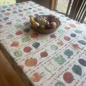 May include: A white tablecloth featuring a repeating pattern of various colorful vegetables including tomatoes, pumpkins, beets, and leafy greens, with a woven brown basket filled with assorted artificial fruits and vegetables placed on top.