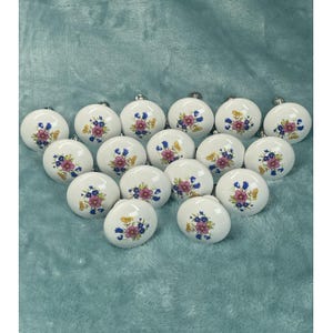 May include: A collection of white ceramic cabinet knobs. Each knob features a floral design with pink, purple, and blue flowers. The knobs are round and have a silver metal base.