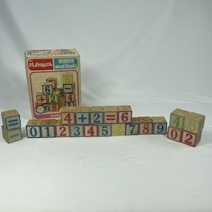 May include: A set of sixteen wooden blocks featuring numbers and mathematical symbols in red, blue, green, and yellow, accompanied by a cardboard box labeled Playskool Number wood blocks, displaying a graphic of the blocks stacked in a tower.
