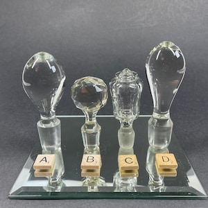 May include: Four clear glass decanter stoppers of varying shapes are arranged in a row. Each stopper has a frosted glass base and is labeled with a small wooden tile marked A1, B3, C3, or D2. The stoppers feature teardrop, faceted, and ornate designs.
