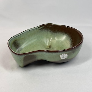 May include: An asymmetrical ceramic bowl with a smooth, glossy finish. The interior features a gradient of sage green and dark brown, while the exterior is primarily sage green with dark brown accents. A small white sticker reads Original Creation by Sankou.