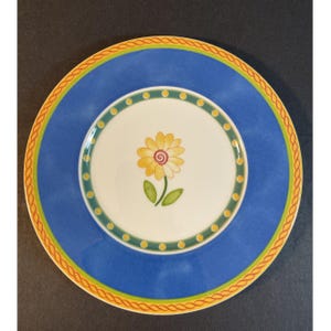 May include: A round ceramic plate with a blue border and a white center. The center features a yellow flower design with green leaves. The plate has a yellow and orange rope-like border.