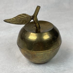 May include: A brass apple-shaped trinket box with a stem and leaf. The box is a warm, golden color, and the leaf and stem are intricately detailed. The lid fits snugly, and the overall design is elegant and decorative.