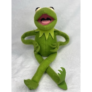 May include: A green felt plush frog character with large white eyes, black pupils, and a wide open mouth showing a pink tongue. The figure has a pointed collar, thin arms with three-fingered hands, and long legs with webbed feet, posed in a seated position.