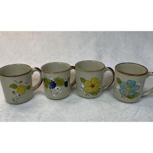 May include: Four ceramic coffee mugs with floral and fruit designs. Each mug has a beige base, brown rim and handle, and a different hand-painted design: lemon, grapes, yellow flower, and blue flower.