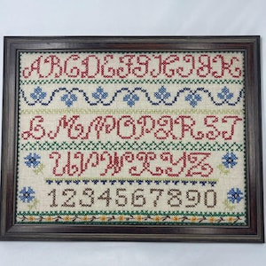 May include: A framed cross-stitch sampler on light beige fabric featuring red ornate capital letters from A to Z, blue floral borders, and brown numbers from 0 to 9. The piece is enclosed in a dark brown wooden frame with a simple rectangular profile.
