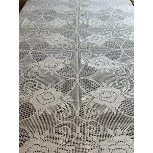 May include: Cream-colored, crocheted tablecloth with a floral pattern. The design features large, openwork roses and geometric shapes, creating a delicate and textured surface. The tablecloth is rectangular and appears to be made of cotton or a similar fiber.