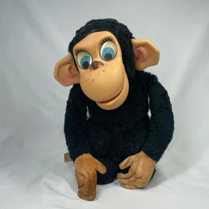 May include: A plush chimpanzee toy featuring a black fuzzy body, large tan ears, and a smooth tan face. The figure has prominent blue eyes with black eyelashes, a small nose, and a light tan mouth area. The hands are made of tan material.