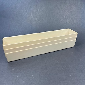 May include: A rectangular, off-white plastic storage bin with an open top and smooth, solid walls. The container features a long, narrow shape with slightly tapered sides and a flat base, designed for organizing items in a drawer or on a shelf.