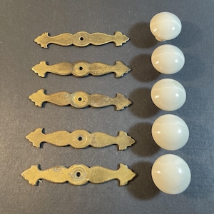 May include: A collection of vintage cabinet hardware, including five brass-toned pulls and five off-white, marble-effect knobs. The pulls have a decorative, elongated design with a central mounting hole. The knobs are spherical.