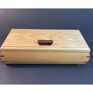 May include: A rectangular wooden box with a light-colored grain lid and a dark brown wooden handle centered on top. The box features curved sides and visible joinery details at the corners, resting on small dark brown feet on a dark surface.