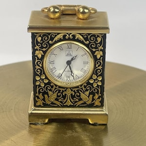 May include: A small rectangular quartz clock with a gold-tone metal top handle and base. The black face features intricate gold-tone floral and bird filigree patterns. The white clock dial displays black Roman numerals and the word QUARTZ.