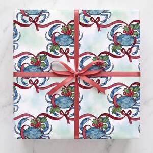 May include: A square gift box wrapped in white paper featuring a repeating watercolor pattern of blue crabs, green holly leaves, and red berries, tied with a solid red satin ribbon that forms a centered bow across the front of the package.