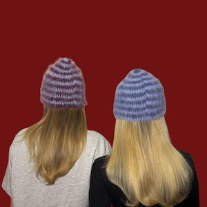 May include: Two individuals are seen from the back, each wearing a knitted beanie. One beanie is a mix of purple and brown stripes, the other is blue. Both have long blonde hair and are set against a red background.