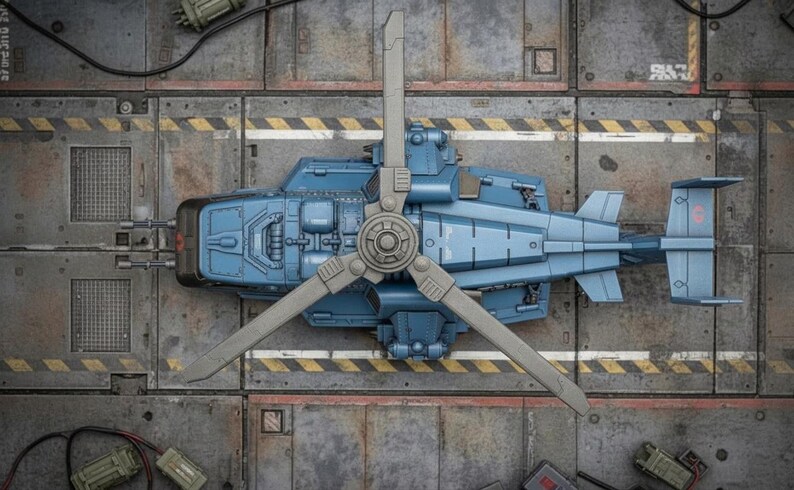 Pu&ograve; includere: Vista aerea dettagliata di un modello di elicottero azzurro con un rotore a tre pale. Il modello &egrave; ambientato su uno sfondo metallico con strisce di pericolo gialle e nere, che suggeriscono un'officina o un hangar.