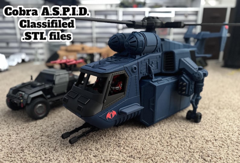 Pu&ograve; includere: Un elicottero giocattolo blu scuro con il testo "Cobra A.S.P.I.D. Classified .STL files" e un logo Cobra rosso. Un camion giocattolo nero &egrave; sullo sfondo. L'elicottero ha un design dettagliato.