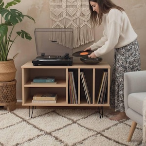 May include: A light wood record player stand with black hairpin legs. The stand holds a record player, records, and a decorative bowl. A woman is placing a record on the player. A large plant and a macrame wall hanging are also in the room.